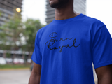 Clear image of royal blue, round neck t-shirt on African American Male standing in front of blurred background scenery of building skyscraper and trees. Printed t-shirt has "Born Royal" in cursive graphic, centered horizontally across chest.  2 lines--Born on first line, Royal on second.  T-shirt 100% cotton.
