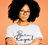 Young, African American woman with full curly afro, and black square eyeglasses, modeling white, round neck Graphic t-shirt. Posing with arms folded. Peach-colored background.  T-shirt graphic says Born Royal- in black, cursive letters, centered across chest in 2 horizontal lines.  