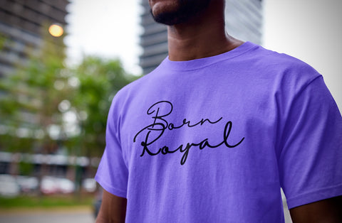 Clear close-up of lavender, round neck t-shirt on African American Male standing in front of blurred background scenery of building skyscraper and trees. Printed t-shirt has "Born Royal" in cursive graphic, centered horizontally across chest.  2 lines--Born on first line, Royal on second.  T-shirt 100% cotton.
