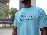 Clear image of light-blue, round neck t-shirt on African American Male standing in front of blurred background scenery of building skyscraper and trees. Printed t-shirt has "Born Royal" in cursive graphic, centered horizontally across chest.  2 lines--Born on first line, Royal on second.  T-shirt 100% cotton.