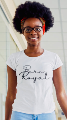 Clear close-up of African American Woman wearing white, round neck Graphic t-shirt standing in a building hallway. Printed t-shirt says "Born Royal" in cursive graphic, centered horizontally across chest.  2 lines--Born on first line, Royal on second.  T-shirt 100% cotton.